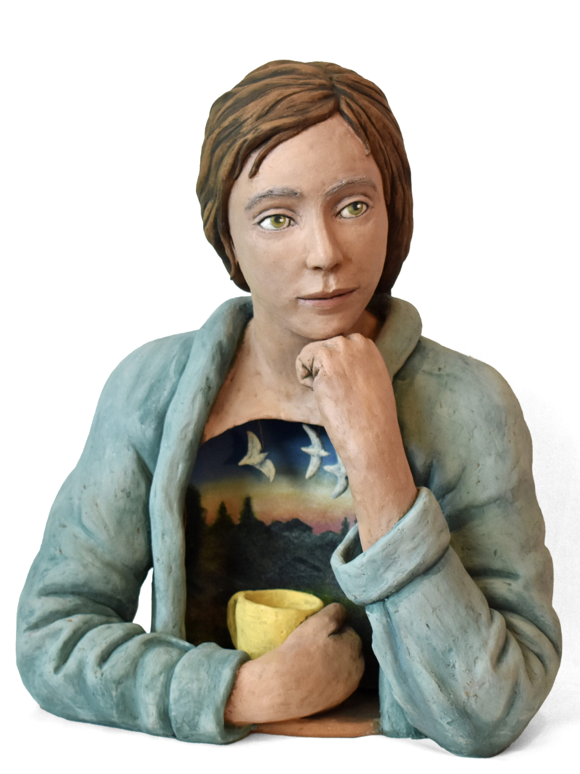A ceramic sculpture of a woman with short cropped hair, her chin resting on her right hand, her left hand clasping a yellow mug. She is wearing a pale blue jacket and a t-shirt with a print of white seagulls flying over pine trees silhouetted against a dawn sky.